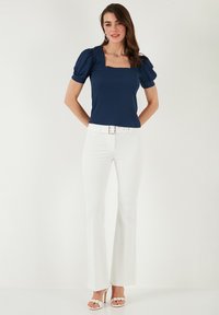 Marineblaues, strukturiertes Oberteil mit Puffärmeln und quadratischem Ausschnitt, kombiniert mit weißen, ausgestellten Hosen und hellen, schlichten Schuhen mit Riemchen.
