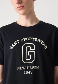 Sort bomulds T-shirt med et stort hvidt G-logo, "GANT SPORTSWEAR" og "NEW HAVEN 1949" tekst i en fed, klassisk skrift.
