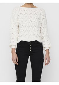 Weißer Strickpullover mit einem strukturierten Rautenmuster und einem weitem Ausschnitt, kombiniert mit schwarzen hochgeschnittenen Jeans mit vier Knöpfen.