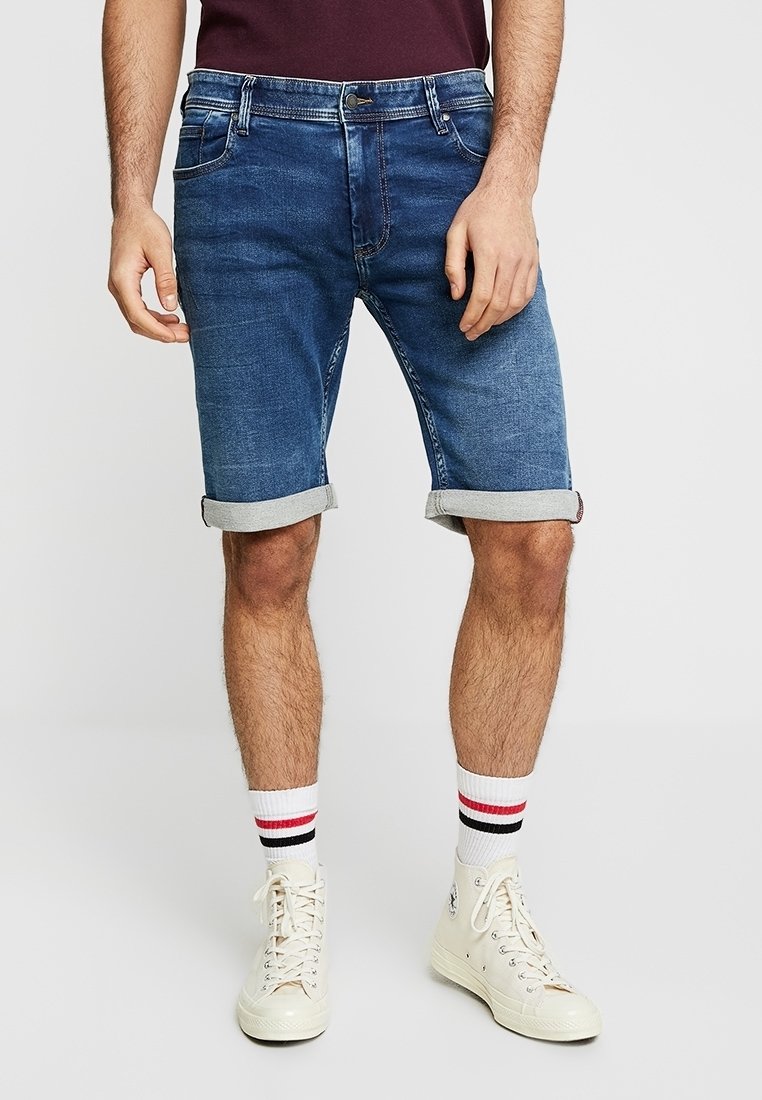 Homme portant un short en jean bleu retroussé, des baskets blanches montantes et des chaussettes blanches à rayures rouges et noires, debout devant un fond uni.