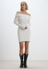 Abito bianco a costine, off-the-shoulder, con maniche lunghe e silhouette aderente, e orlo sopra il ginocchio. Abbinato a stivali cowboy neri.