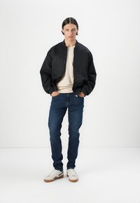 Chaqueta bomber negra con cremallera, llevada sobre una camisa de color claro. Combinada con jeans azul oscuro y zapatillas blancas con acentos marrones.