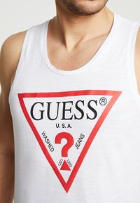 Débardeur blanc présentant un logo triangulaire rouge de Guess avec les éléments de texte "WASHED", "JEANS" et un point d'interrogation rouge au centre.