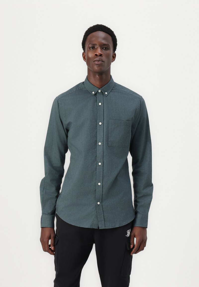 Camicia verde scuro a maniche lunghe con colletto a punta; presenta una tasca sul petto e bottoni bianchi; texture liscia e tessuta.