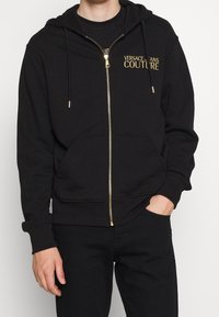 Sudadera con cremallera negra hecha de material suave. Presenta un logo dorado de "Versace Jeans Couture" en el pecho, cordones y bolsillos frontales.