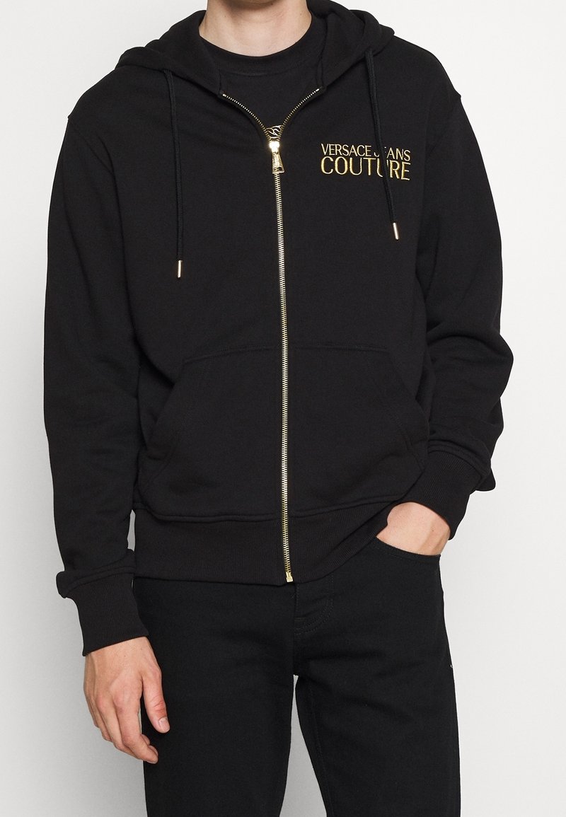Sudadera con cremallera negra hecha de material suave. Presenta un logo dorado de "Versace Jeans Couture" en el pecho, cordones y bolsillos frontales.
