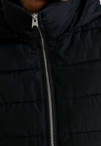 Sort polstret jakke med høj krave, der har en sølvfarvet lynlås og vandret syninger. Materialet virker glat og vejrbestandigt.