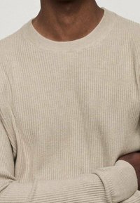 Ein dunkelhäutiger Mensch trägt einen beigen, gerippten Pullover mit langen Ärmeln und rundem Halsausschnitt, die Arme am Bauch verschränkt.