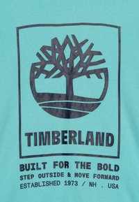 Tessuto turchese con un logo di un albero nero su una base circolare. Il testo include "TIMBERLAND" e "BUILT FOR THE BOLD" in carattere grassetto.
