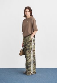 Tricou mare, de culoare maro, cu un logo, asociat cu pantaloni evazați în camuflaj verde, cu dungi albe. Accesoriu: o geantă mică, bej.