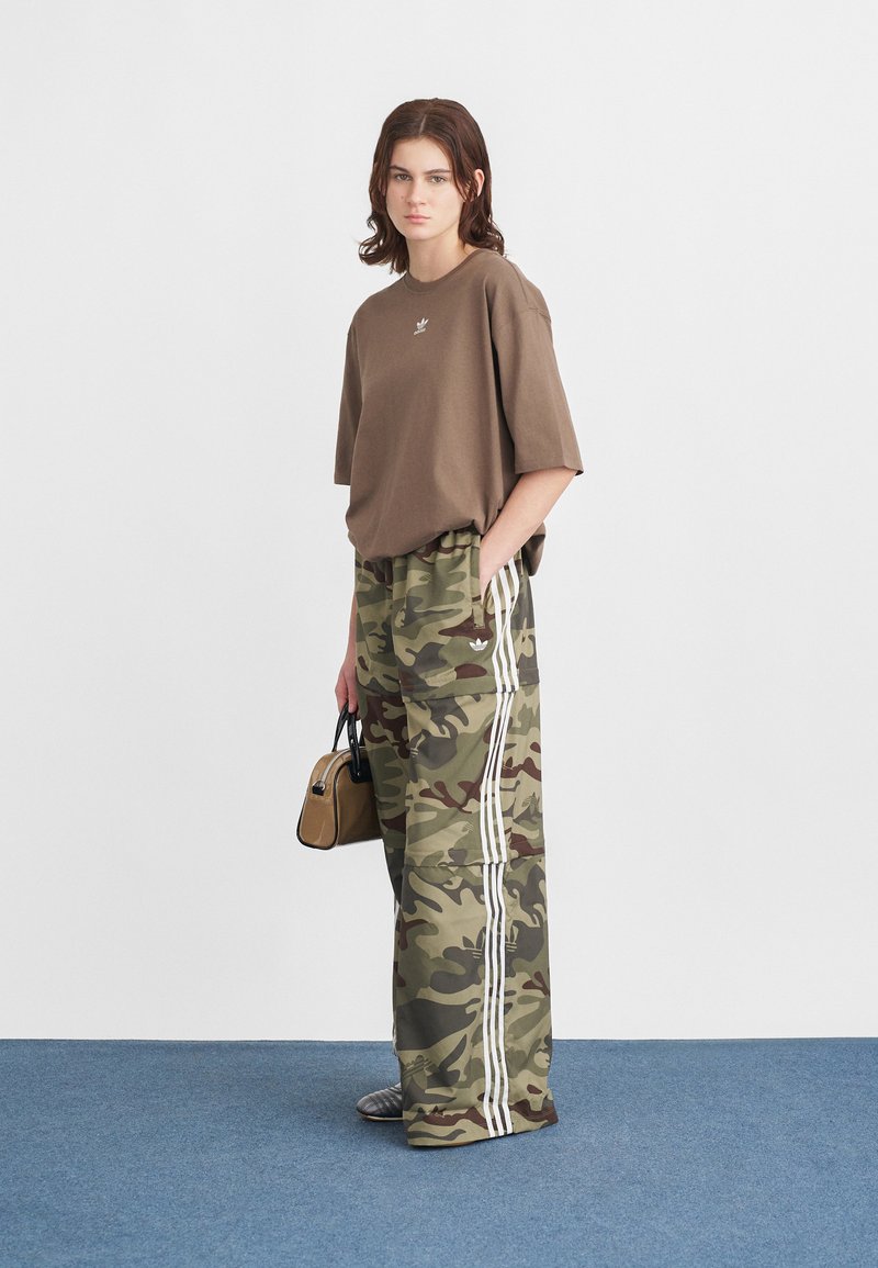 Braunes oversized T-Shirt mit einem Logo, kombiniert mit weiten grünen Camo-Hosen mit weißen Streifen. Accessoire: kleine beige Handtasche.