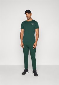 Mørkegrønt treningsantrekk bestående av en tettsittende kortermet t-skjorte og tapered joggers. Svart caps med logo, kombinert med svarte sneakers.