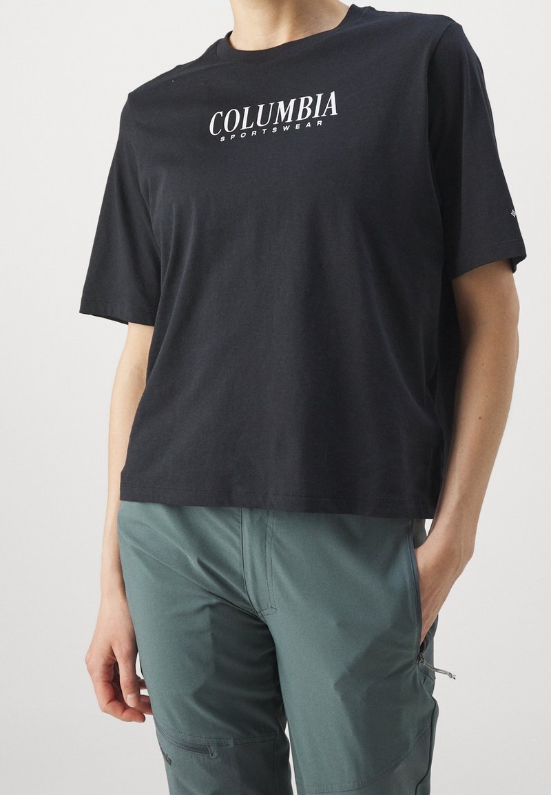 Svart kortärmad t-shirt med "COLUMBIA"-logotyp; mjukt bomullsmaterial, något oversized passform, rundad halsringning, tillsammans med gröna byxor.