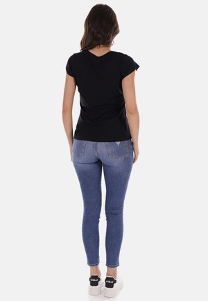 T-shirt nera a maniche corte, taglio aderente, abbinata a jeans skinny blu chiaro. Indossando sneakers bianche con dettagli neri. Vista da dietro.