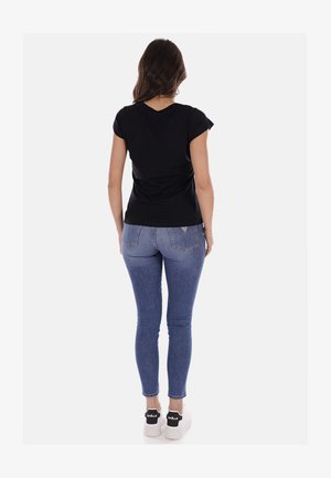 T-shirt nera a maniche corte, taglio aderente, abbinata a jeans skinny blu chiaro. Indossando sneakers bianche con dettagli neri. Vista da dietro.