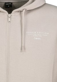 Helle graue Zip-Hoodie aus weichem Stoff, mit einer Kapuze mit Kordelzug und einer Fronttasche. Text auf der Brust in weißer Schrift.