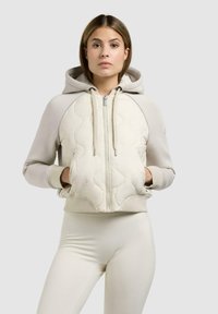 Kvinde iført lys beige quiltet hættetrøje med lynlås og lommer samt matchende leggings, stående mod en ensfarvet grå baggrund.