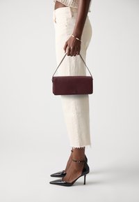 Bordeaux leren clutch met een gestructureerde afwerking en een dunne schouderband, vastgehouden door een persoon in lichtgekleurde, cropped broek en zwarte hakken.
