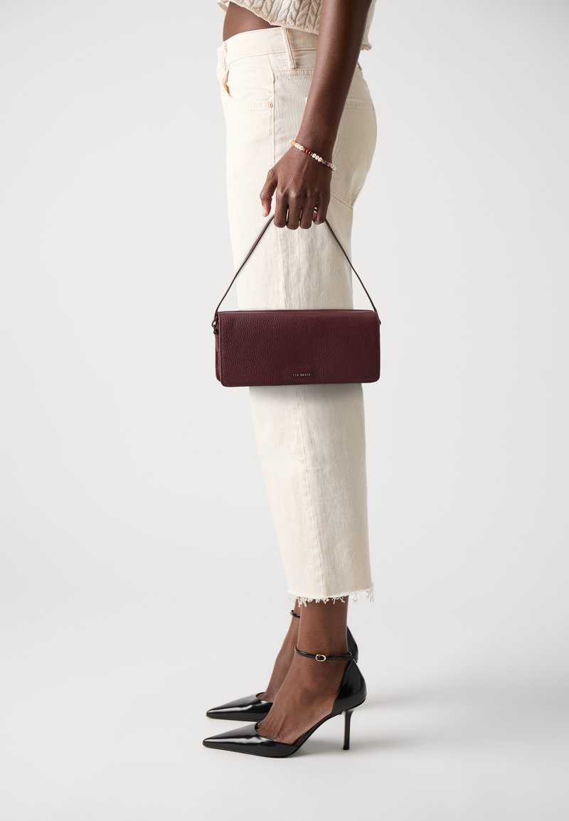 Bordeaux leren clutch met een gestructureerde afwerking en een dunne schouderband, vastgehouden door een persoon in lichtgekleurde, cropped broek en zwarte hakken.