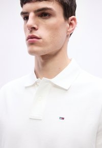 Νέος άνδρας με κοντά καστανά μαλλιά που φοράει λευκό πόλο πουκάμισο με γιακά και ένα μικρό κόκκινο, λευκό και μπλε λογότυπο στο στήθος.