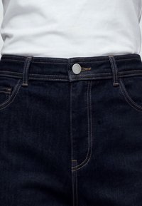 Jean en denim bleu foncé avec coutures marron et bouton argenté porté avec une chemise blanche rentrée.