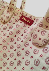 Borsa tote in tessuto beige chiaro con motivi ripetuti di 'P' bordeaux e alloro, e un'etichetta in pelle bordeaux con il nome "Pollini".