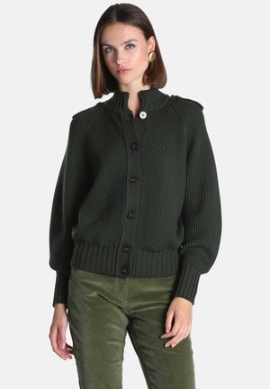 Femme portant un cardigan tricoté côtelé vert foncé avec de gros boutons et un pantalon vert assorti, debout sur un fond uni.