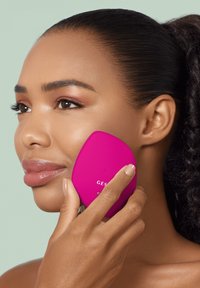 Appareil de nettoyage facial en silicone de couleur rose vif, en forme ovale avec des boutons pour les réglages, tenu contre la joue d'un modèle avec une peau lisse.