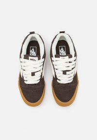 Sapatilhas de camurça castanha com atacadores brancos, uma faixa lateral branca e uma sola de borracha de goma. Apresenta um colarinho acolchoado e o logo da Vans na língua.