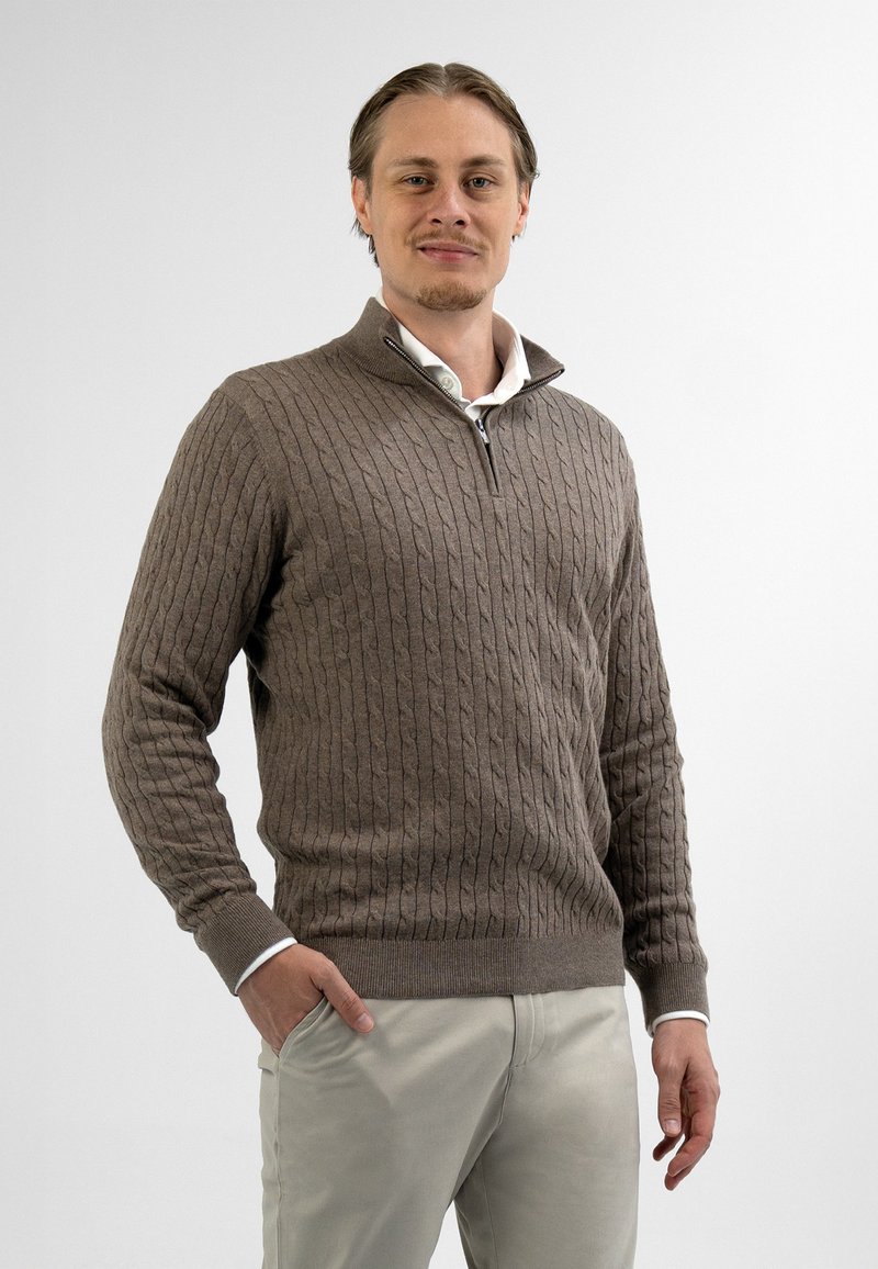 Bruine gebreide trui met een ritskraag, voorzien van een gestructureerd kabelpatroon. Draagt over een witte overhemd met kraag, gecombineerd met lichtbeige broek.