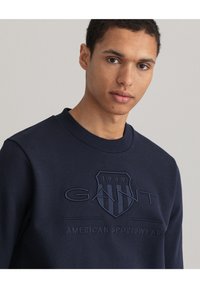 Mörkblå sweatshirt med en texturerad, broderad logotyp som visar "GANT" och "AMERICAN SPORTSWEAR", rund halsringning och ribbade ärmslut.