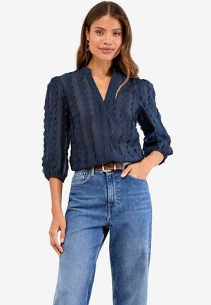 Marinebluse mit V-Ausschnitt, strukturiertem Design mit gewellten Linien und Dreiviertel-Puffärmeln, kombiniert mit hellblauen Jeans.