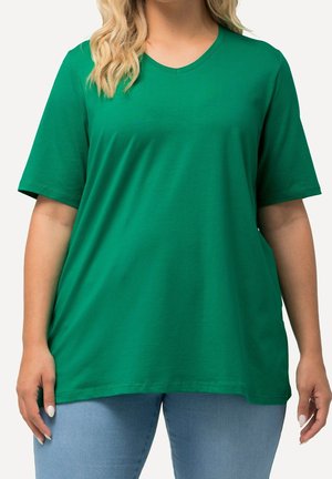 Osoba nosząca jednolity zielony t-shirt z krótkim rękawem i dekoltem w serek oraz jasnoniebieskie dżinsy, widoczna od ramion do bioder na białym tle.