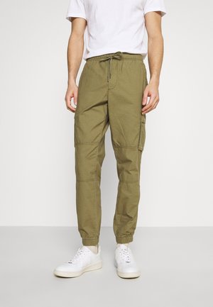 Mand iført olivengrønne cargobukser, hvide sneakers og en hvid t-shirt med korte ærmer, står foran en ensfarvet lys baggrund.