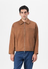 Brun mockaskinnjacka med krage, framdragkedja och sidofickor. Har en avslappnad passform och en mjuk yta. Buren över blå denim.