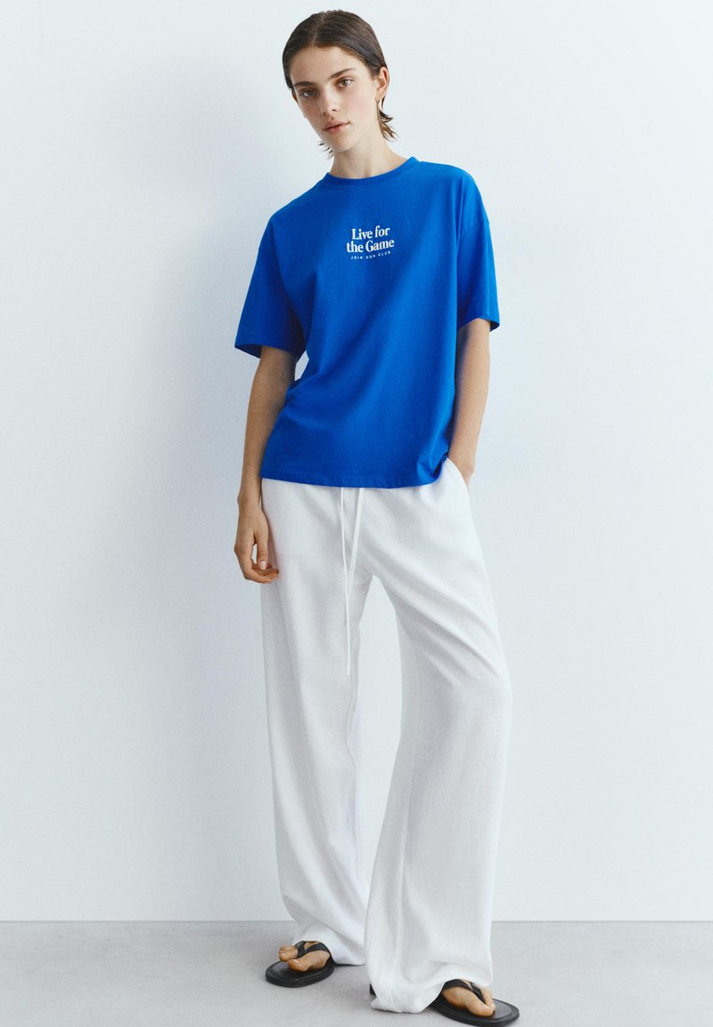 Jeune personne debout, portant un T-shirt bleu avec le texte « Live for the Game », un pantalon large blanc et des tongs noires devant un mur blanc.