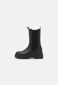 Bota de cuero negra hasta la mitad de la pantorrilla con suela gruesa, panel lateral elástico, tiradores y pequeño logo dorado "Tommy Hilfiger" cerca del talón.