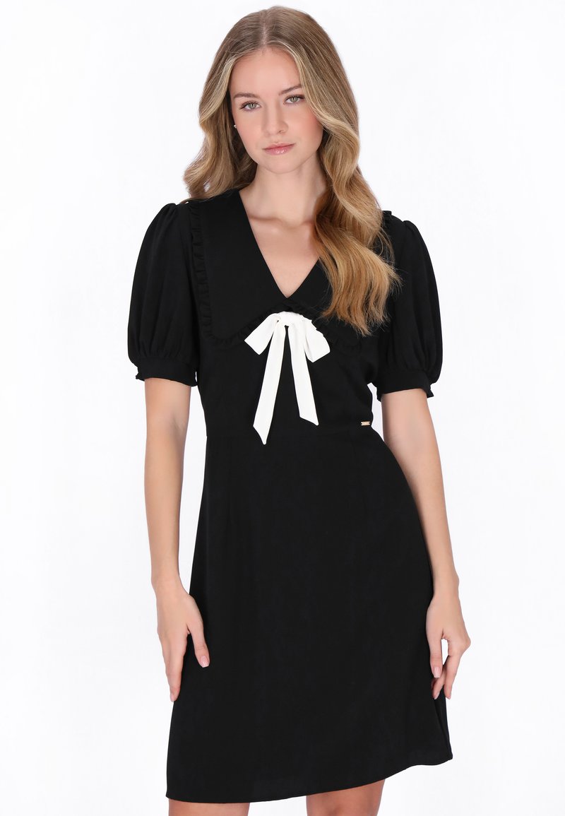 Jonge vrouw met lang golvend blond haar, gekleed in een zwarte jurk met pofmouwen en een witte strik bij de halslijn, staand tegen een witte achtergrond.