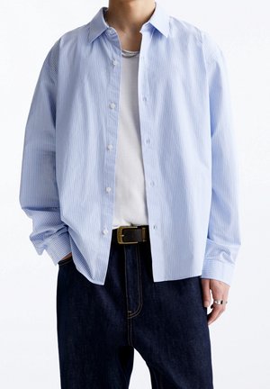 Persoon draagt een lichtblauw-wit gestreept overhemd met verticale strepen over een wit T-shirt, een donkere spijkerbroek met een zwarte riem en een zilveren ketting en ringen.