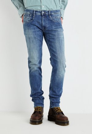Jean en denim bleu avec une coupe slim, présentant des détails de délavage, cinq poches et une fermeture à bouton, associés à des bottes en cuir marron à lacets.