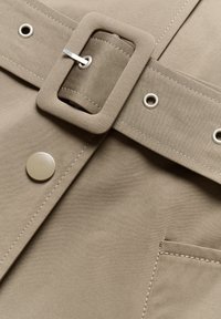 Cintura in tessuto beige con una grande fibbia quadrata e dettagli in metallo argentato, dotata di un bottone a pressione e cuciture visibili su una tasca.
