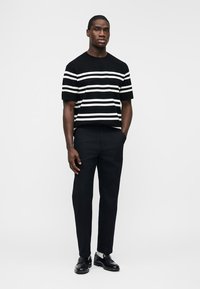 Mann trägt schwarzen Kurzarmpullover mit weißen horizontalen Streifen, schwarze Hose, weiße Socken und schwarze Schnürschuhe, steht mit Händen in den Taschen.