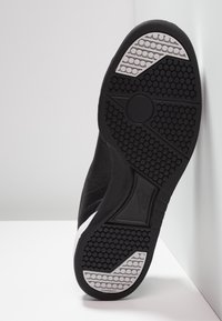 Suela de zapatilla negra con superficie de goma texturizada, que presenta patrones hexagonales y un acento blanco a lo largo del borde. Área del talón reforzada visible.