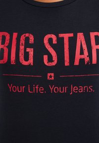 Raudonos nusidėvėjusios „BIG STAR“ raidės virš šūkio „Jūsų gyvenimas. Jūsų džinsai.“ ant juodo audinio fono.