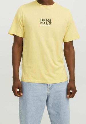 Hombre que lleva una camiseta de manga corta de color amarillo pálido con texto en negro y blanco que dice "ORIGINALS WORLDWIDE", combinada con vaqueros azul claro.