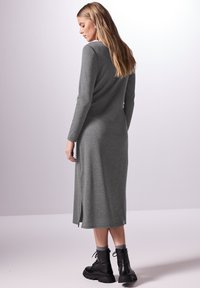 Abito lungo in maglia grigia a maniche lunghe con spacco laterale, abbinato a stivaletti neri alla caviglia. Texture liscia, silhouette aderente e design minimalista.