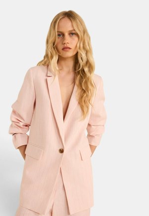Femme aux longs cheveux blonds ondulés portant un blazer rose clair à un bouton et un pantalon assorti, debout sur un fond uni.