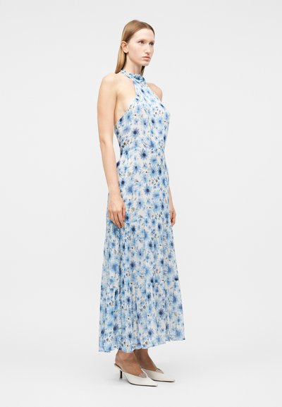 Γυναίκα που στέκεται με ένα αμάνικο, μακρύ φόρεμα halter με μπλε και λευκά λουλούδια και λευκά μυτερά τακούνια μπροστά σε απλό φόντο.