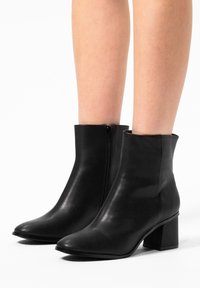 Zwarte leren enkelbootjes met een vierkante neus en blokhak. Voorzien van een zijrits en een gladde textuur; minimalistisch ontwerp.