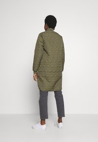 Olivgrön quiltad långjacka med hög krage, med mönster av diamantformer, sidofickor och långa ärmar. Bärs med grå jeans och vita skor.